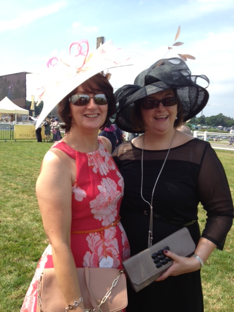 Two friends at the races wearing wide-brimmed occasion hats, one in soft pink florals and one in black sheer with feathers