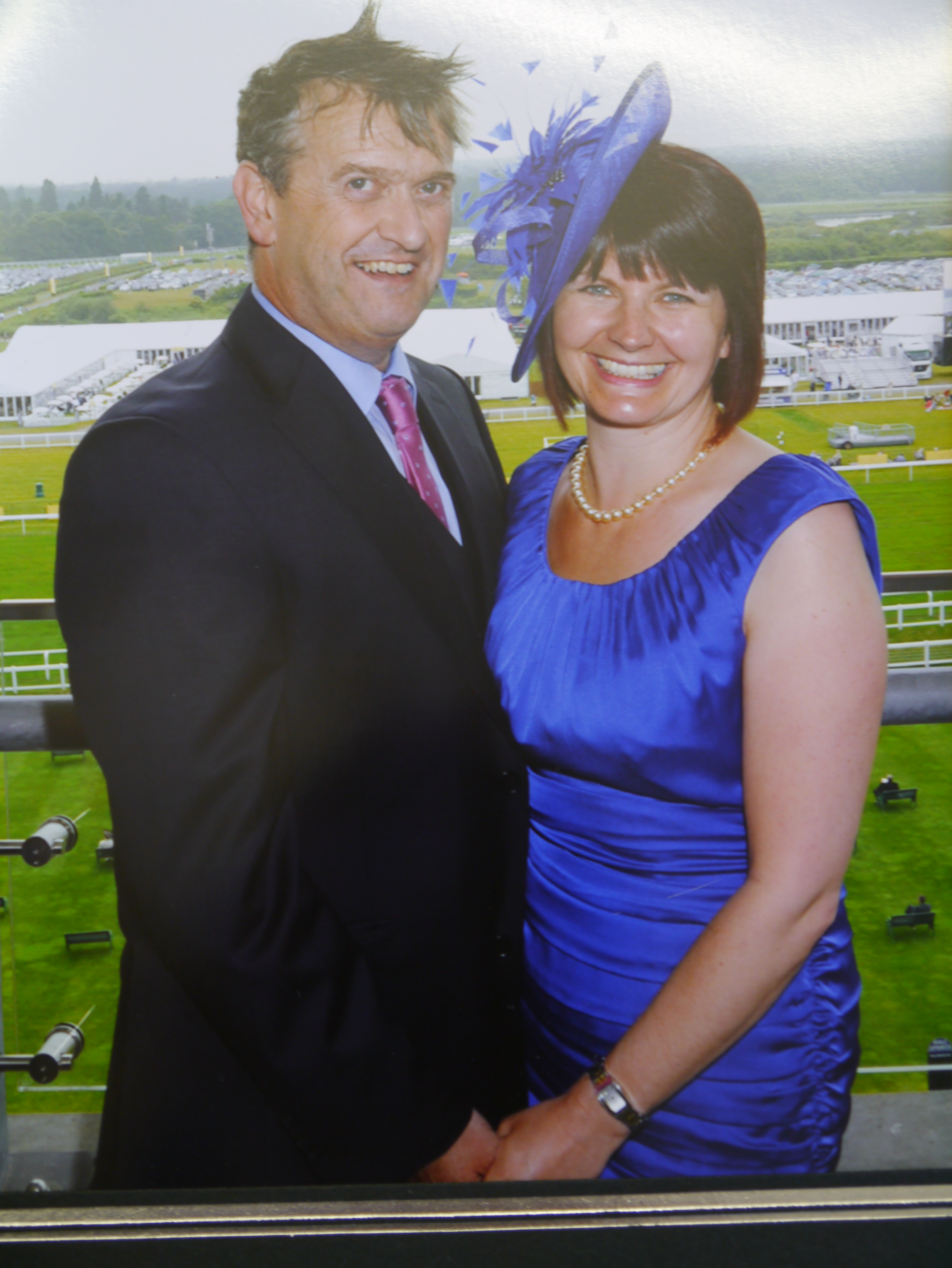 Race day guest in a royal blue dress wearing a bold blue and purple feathered fascinator, overlooking the racecourse
