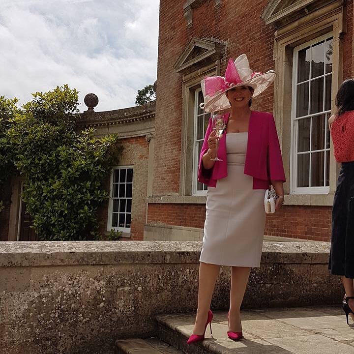Guest in a fuchsia jacket and blush, feather-trimmed hat, enjoying a celebration in the gardens