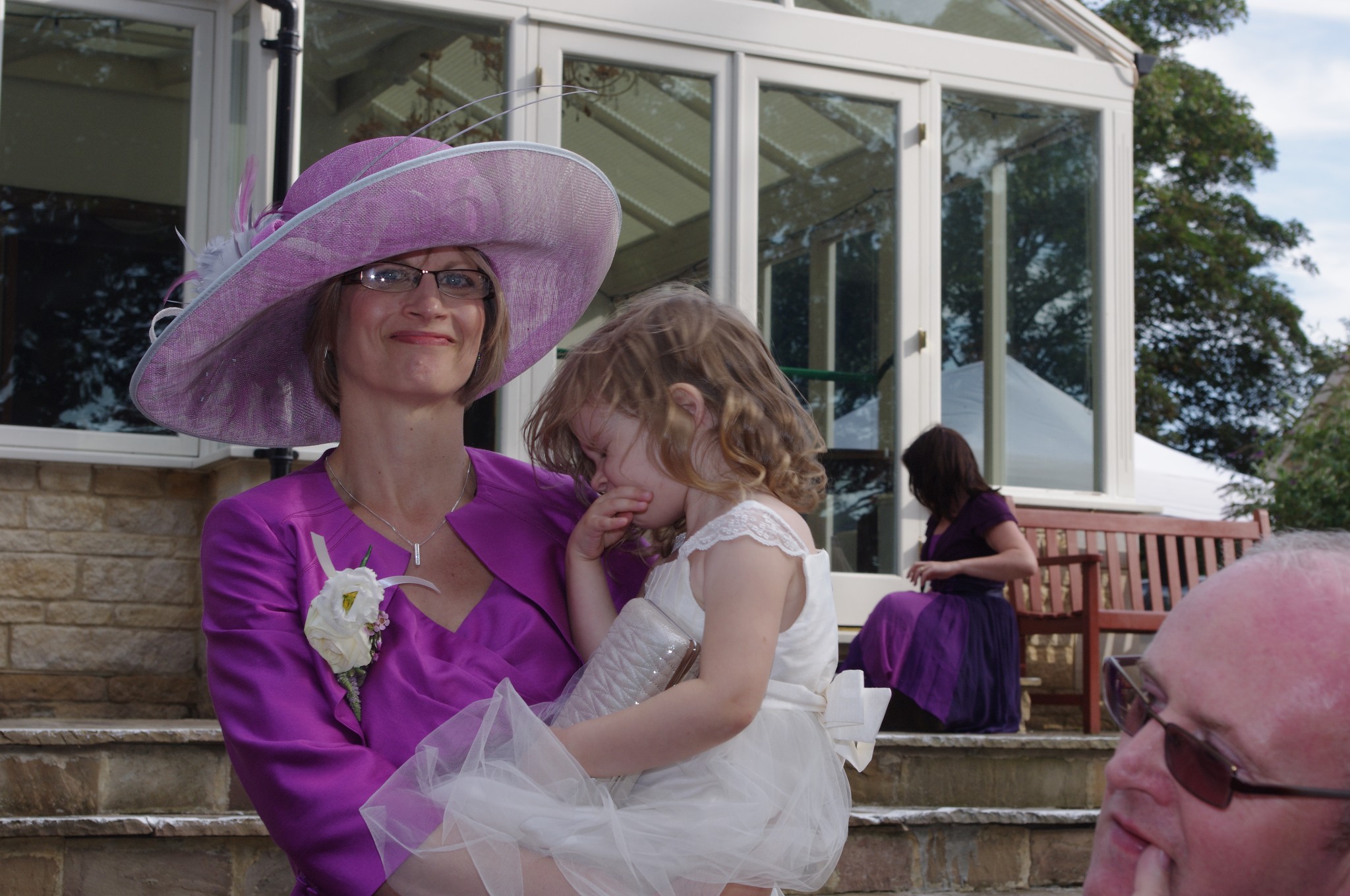Mother of the bride wearing a purple wide-brimmed hat with feather trim and a matching jacket, holding a flower girl